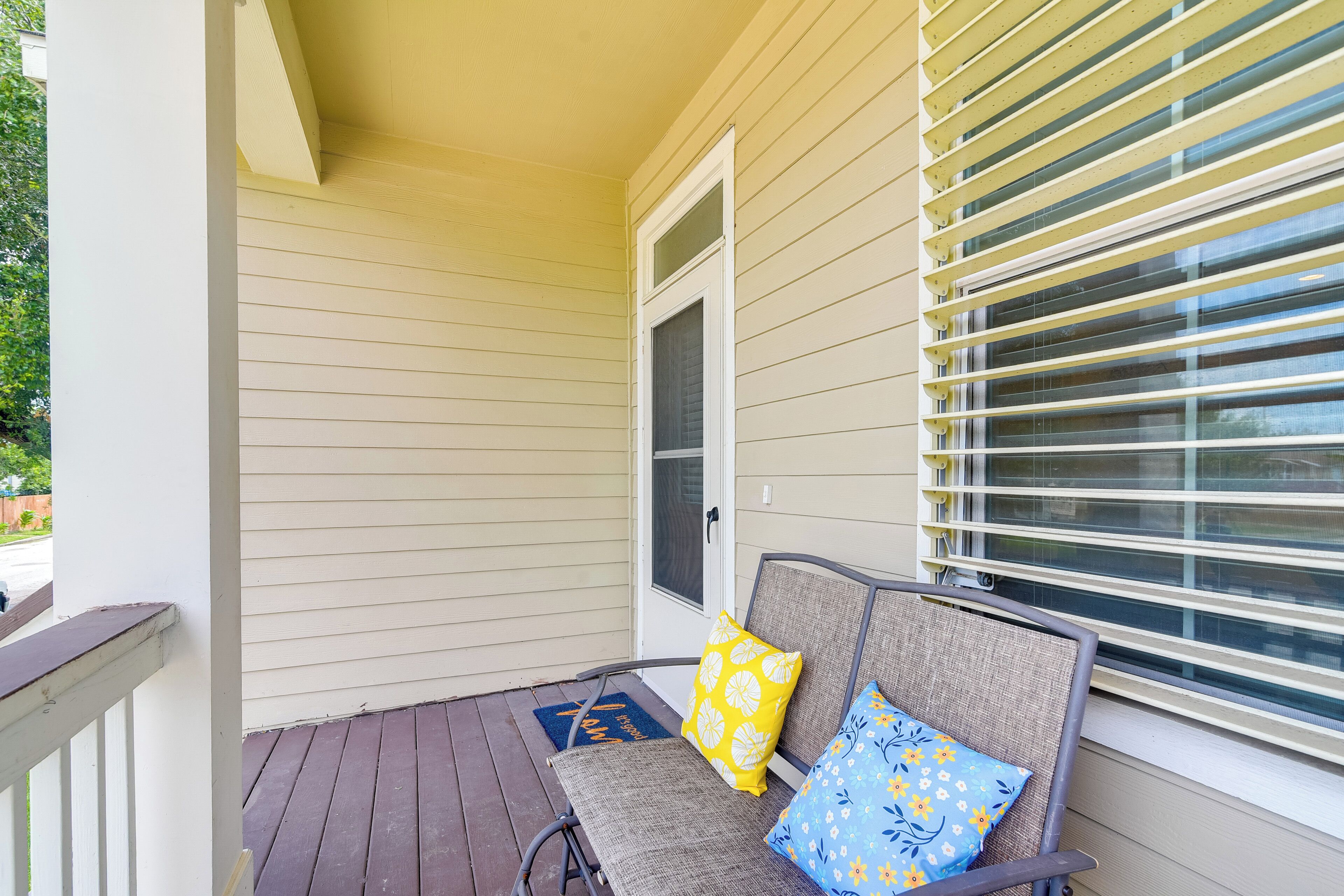 A covered porch with a bench and two pillows, a door, and a window with blinds.