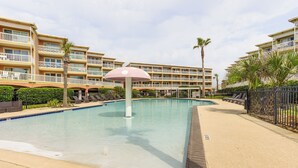 Una piscina con un paraguas rosa, rodeada por una cerca y palmeras, con un edificio de hotel de varios pisos al fondo.