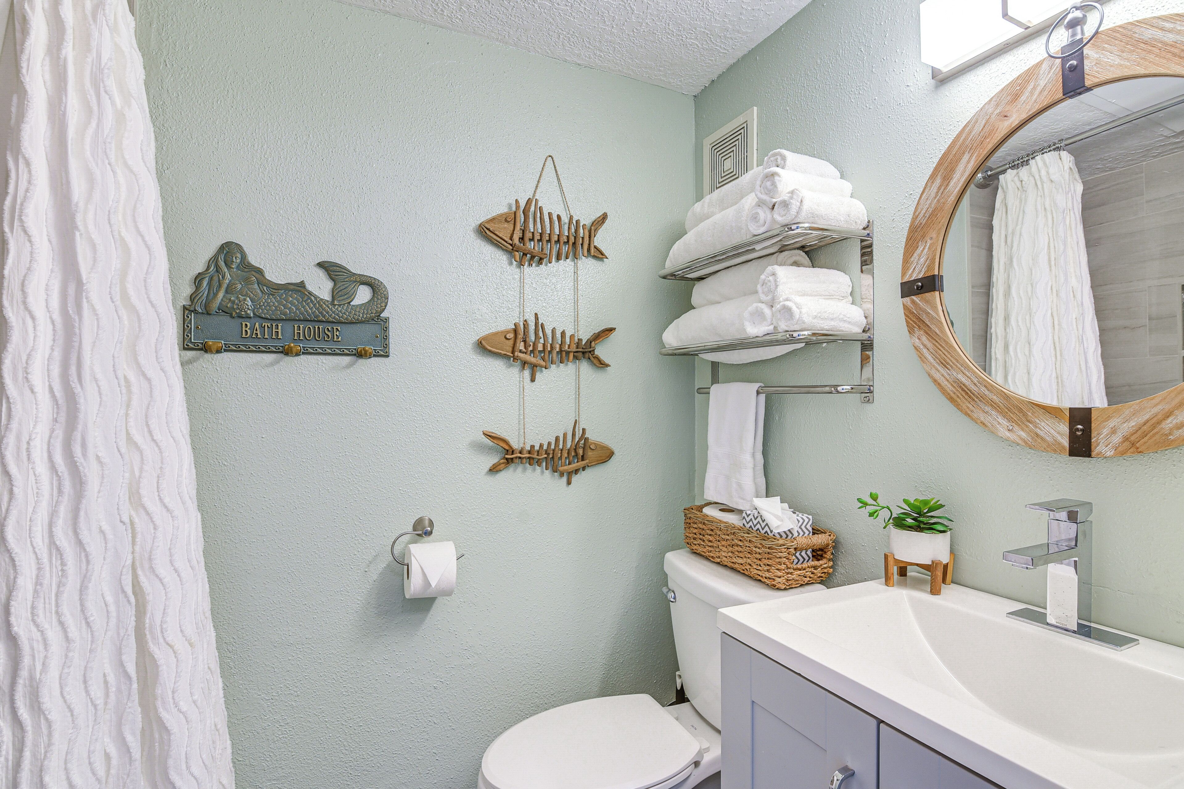 Un bagno con un water bianco, un lavandino con uno specchio, asciugamani impilati su uno scaffale e un quadro decorativo con soggetti marini.