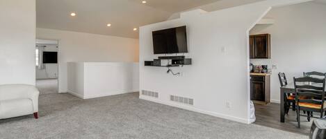 A spacious living room with a flat-screen TV mounted on the wall, a dining area with a table and chairs, and a kitchen area with wooden cabinets.