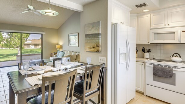 A modern kitchen with white cabinets, a refrigerator, and a microwave. There is a dining table with chairs and a ceiling fan.