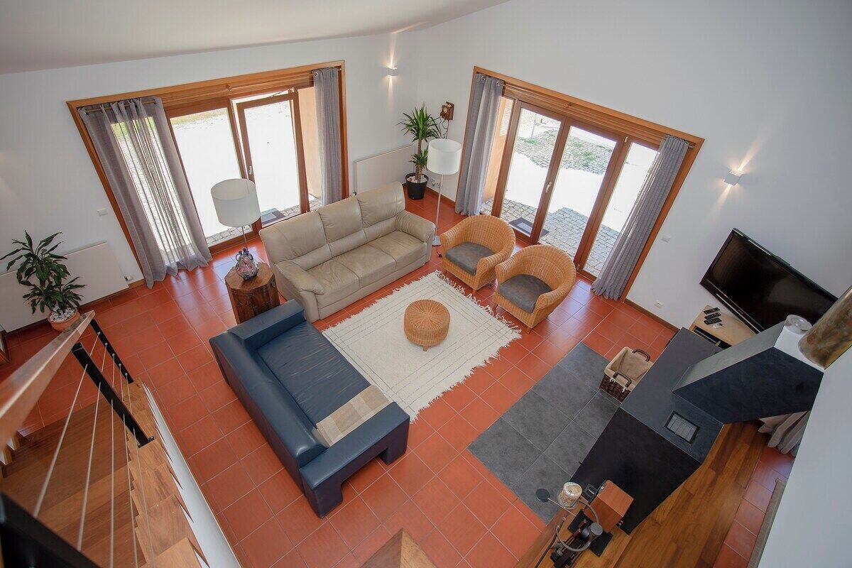 Ein geräumiger Wohnbereich mit einer Couch, Sesseln, einem Couchtisch und einem Fernseher. Große Fenster mit Vorhängen, eine Treppe und ein Blumentopf sind ebenfalls vorhanden.