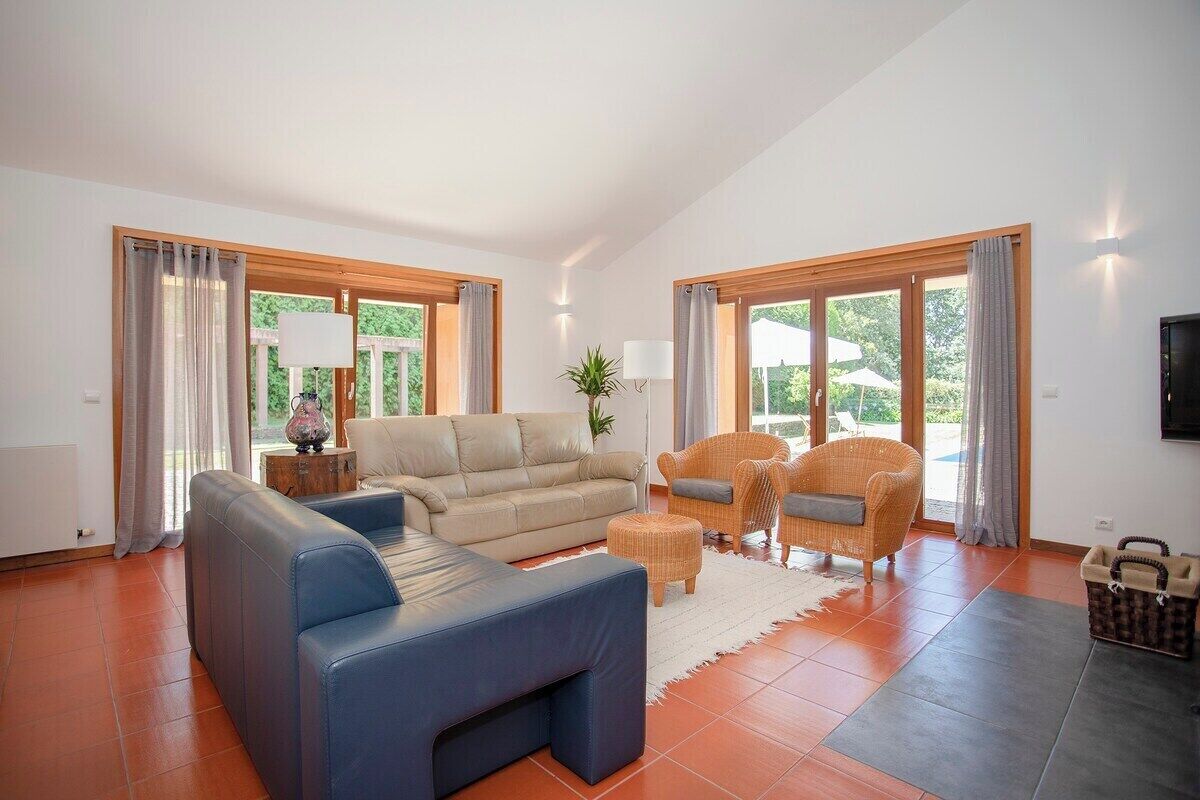 Un salon spacieux avec un canapé, des fauteuils et une table basse. De grandes fenêtres sont agrémentées de rideaux, et on y trouve également un téléviseur et une plante.