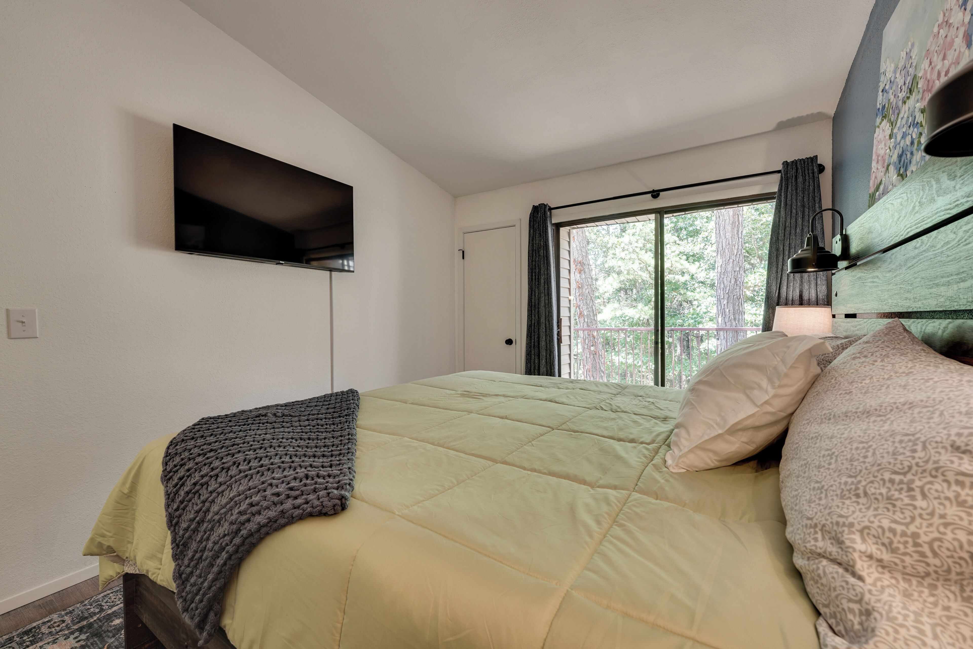 Ein Schlafzimmer mit einem Bett, einem an der Wand montierten Fernseher, einem Fenster mit Vorhängen und einer Lampe auf einer Holzkommode.