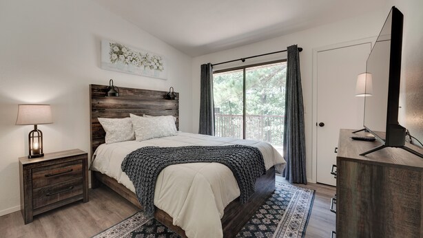 A bedroom with a wooden headboard, a bed with white bedding, a nightstand with a lamp, a large window with curtains, and a TV on a wooden stand. - 0.5 Mi to Table Rock Lake: Ozark Mountains Cabin (Kimberling City)