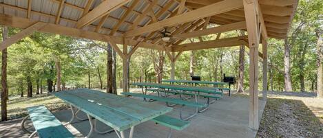 A covered picnic area with multiple picnic tables and benches, surrounded by trees.