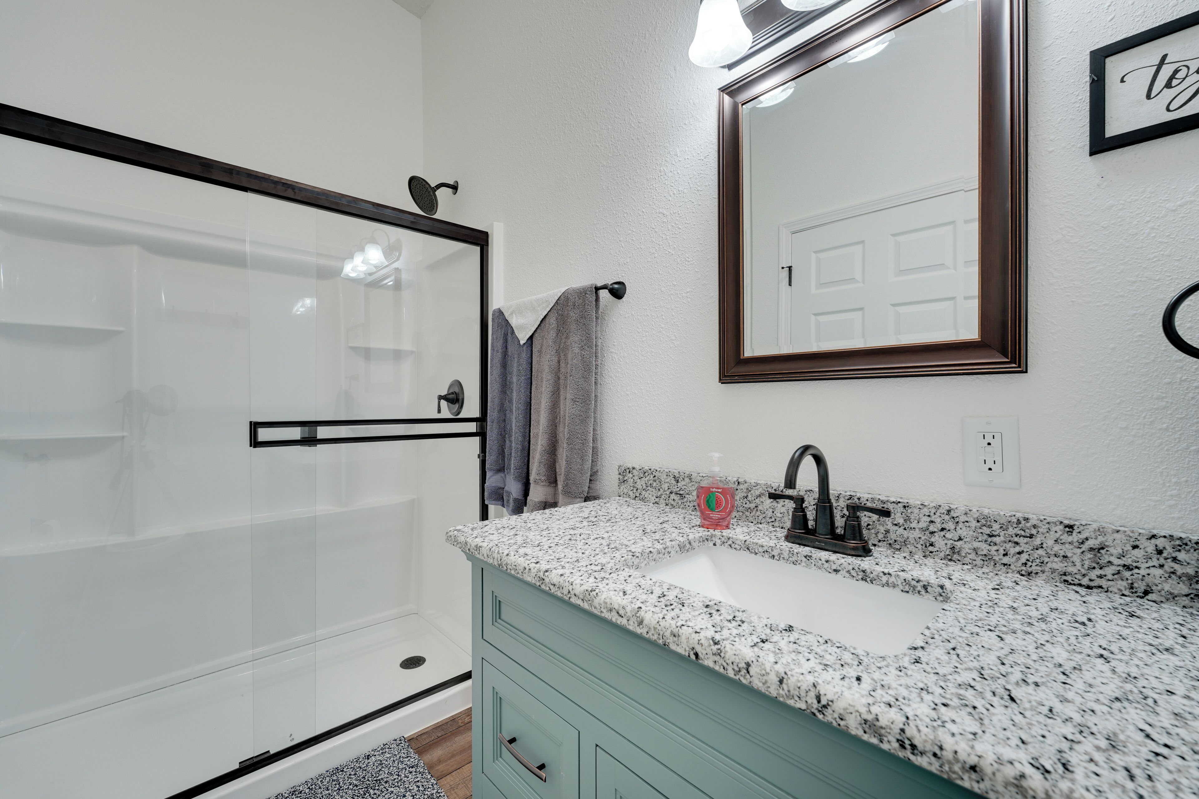 A bathroom with a glass shower door, a sink with a granite countertop, and a mirror with a 'Together' sign.