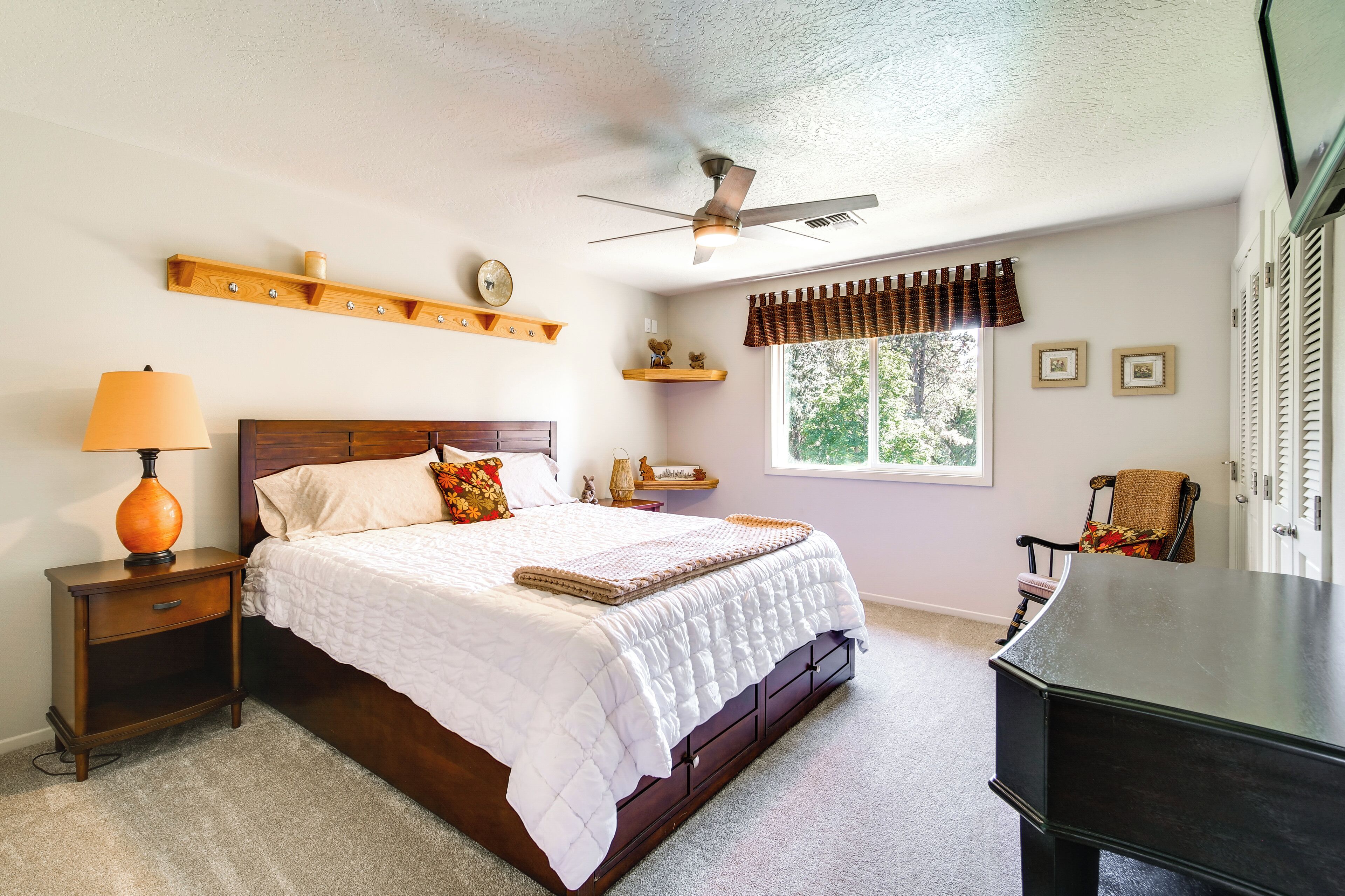 A bedroom with a bed, nightstand, lamp, ceiling fan, window with curtains, and a dresser.