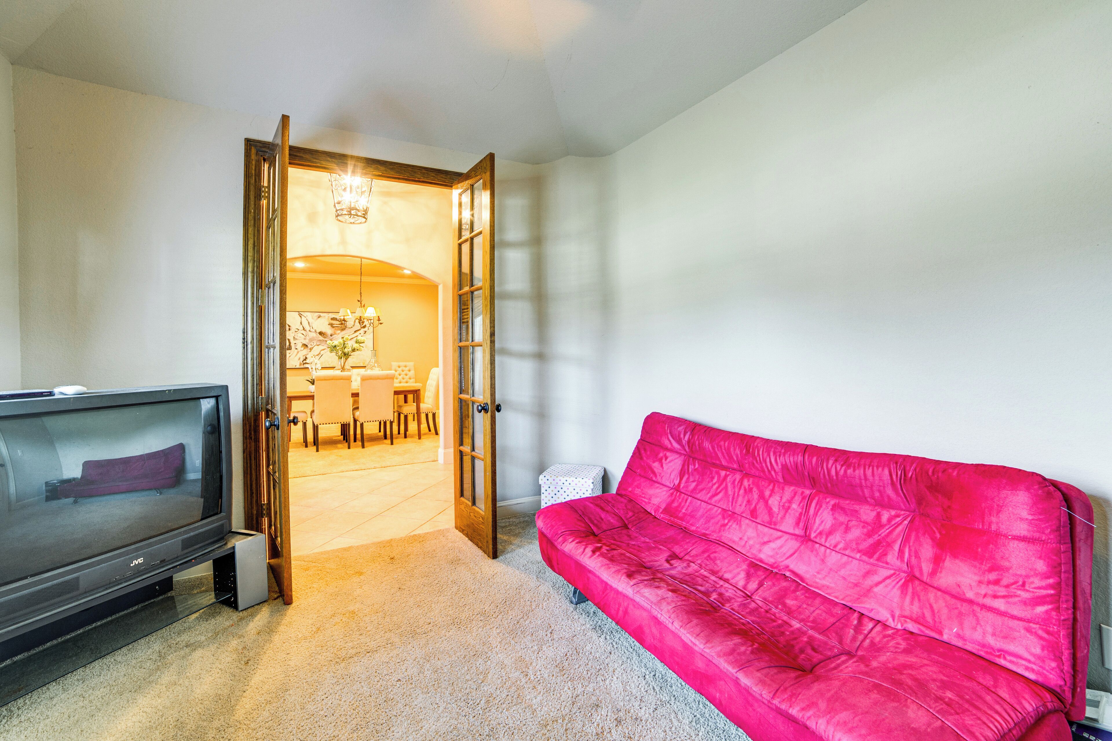 A living room with a TV, a red sofa, and an open door leading to a dining area.