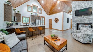 A spacious living area with a wooden ceiling, a stone fireplace, and a kitchen with a large island and modern appliances. - Fire Pit & Hot Tub: Couples Retreat in Broken Bow (Broken Bow)