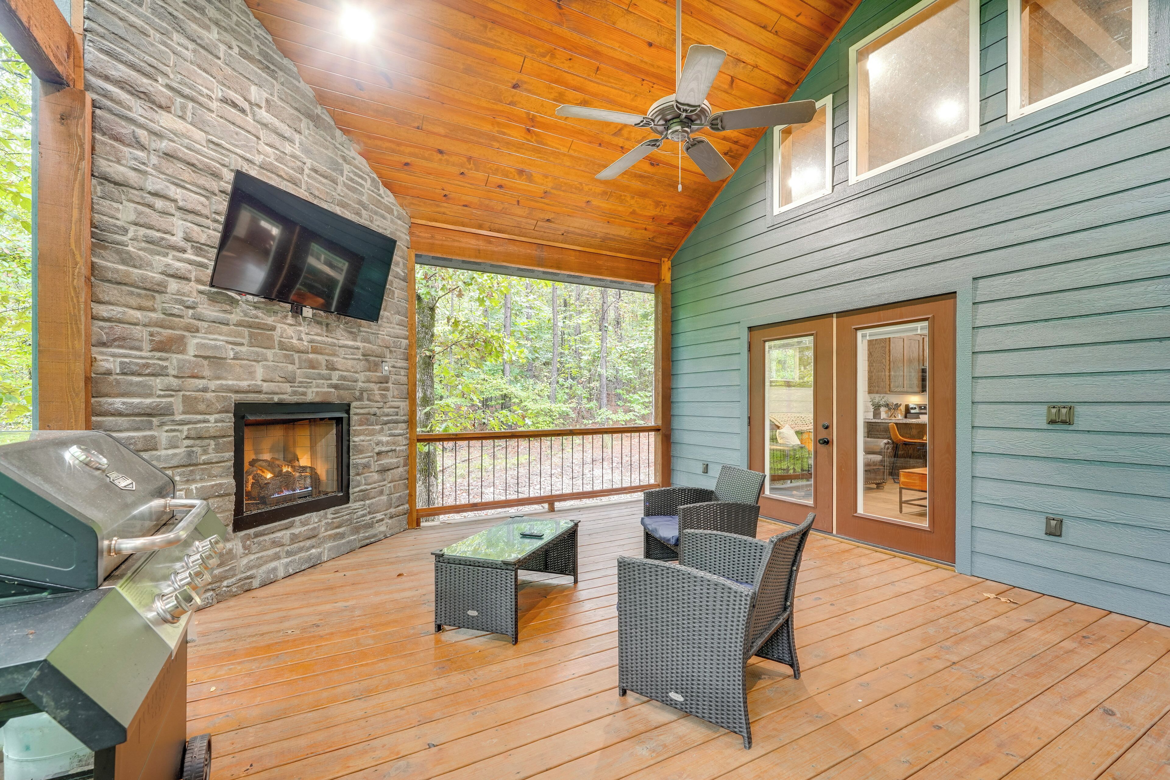 A covered outdoor area with a stone wall, a TV mounted on it, a grill, wicker furniture, and a ceiling fan.
