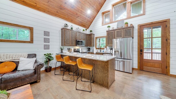A modern kitchen with wooden cabinets, a large island with bar stools, a refrigerator, and a microwave. - Fire Pit & Hot Tub: Couples Retreat in Broken Bow (Broken Bow)