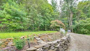 A stone wall along a gravel path in a forested area with green grass and trees. - Deck, Fire Pit, Games: Family Escape Near Boone (Zionville)