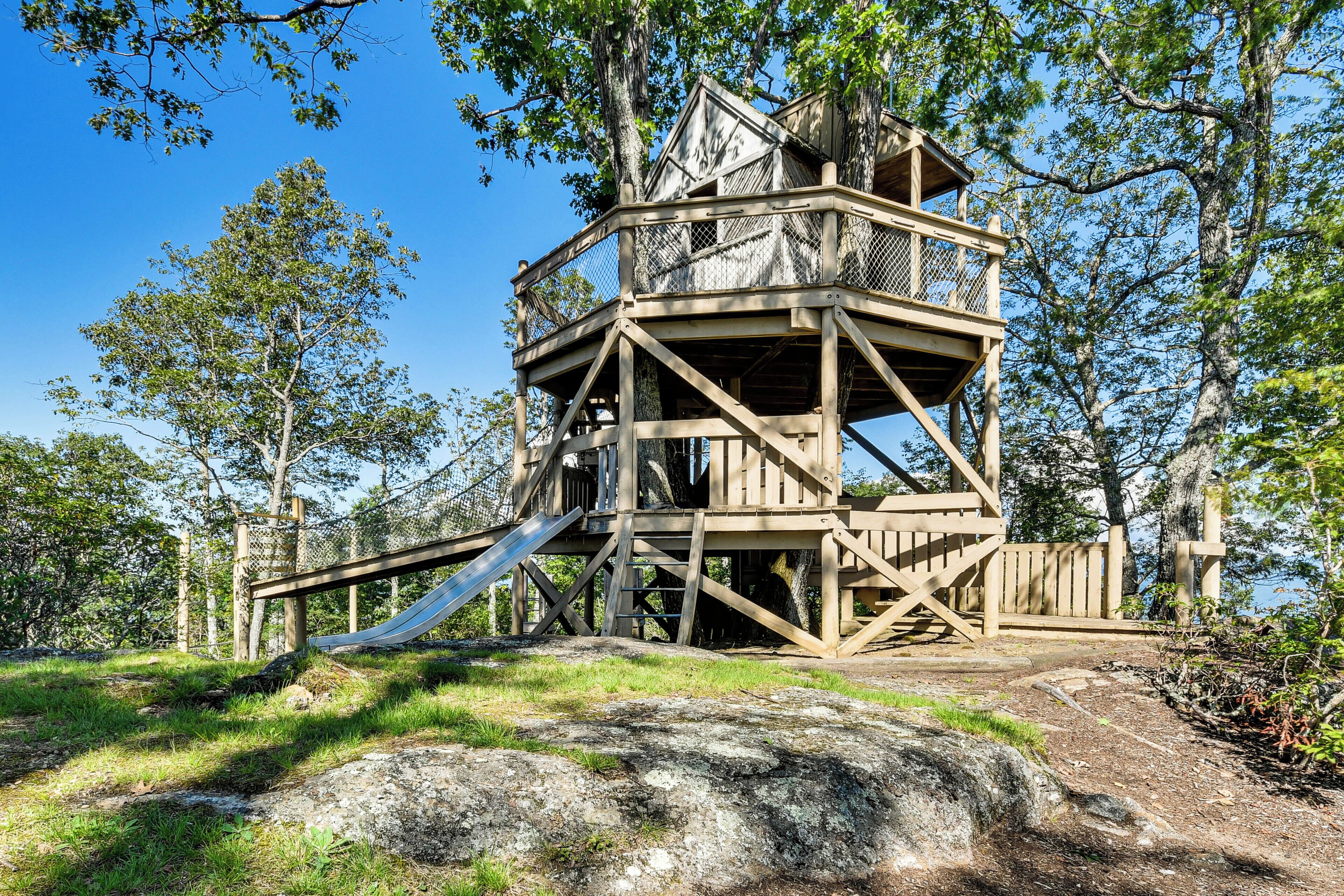 A wooden observation tower with a slide, surrounded by trees and a clear sky.