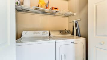 A laundry room with a front-loading washer and dryer, a shelf with various items, and a door to the right.