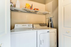 Una sala de lavandería con lavadora y secadora de carga frontal, un estante con varios objetos, y una puerta a la derecha.