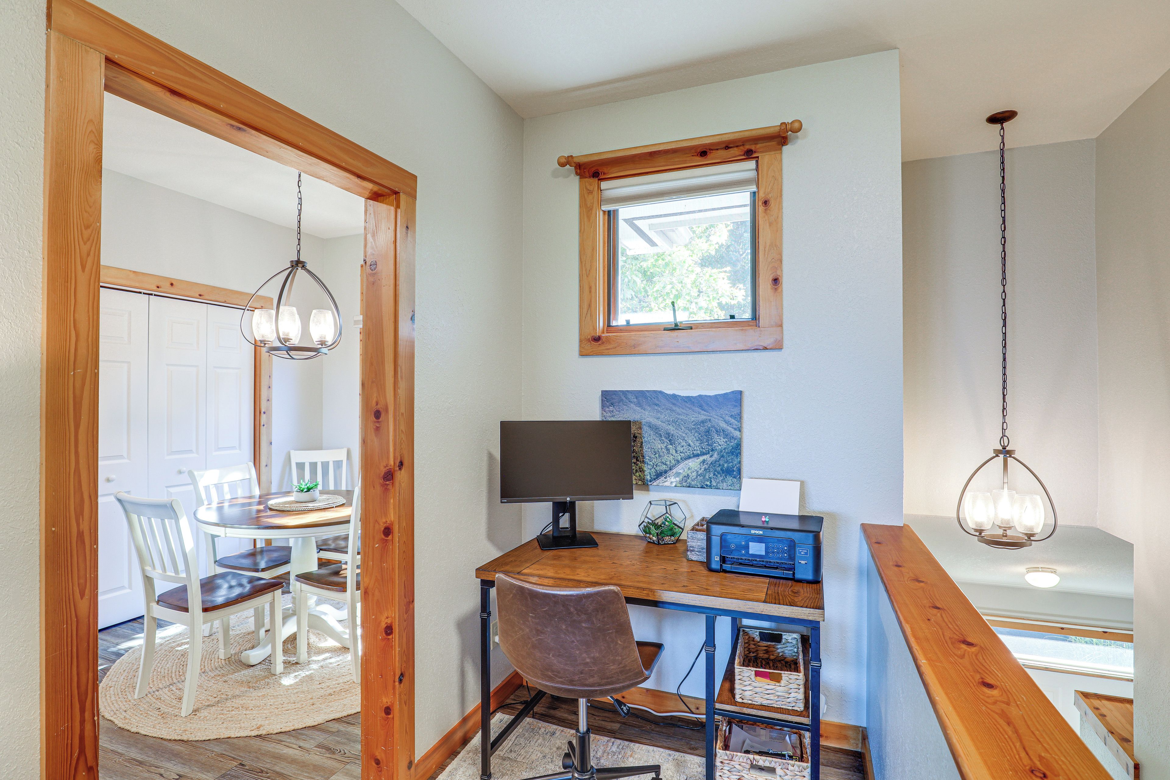 Una habitación con escritorio de madera, computadora, impresora y una silla. Hay una ventana con vista a árboles y una puerta que da a otra habitación.