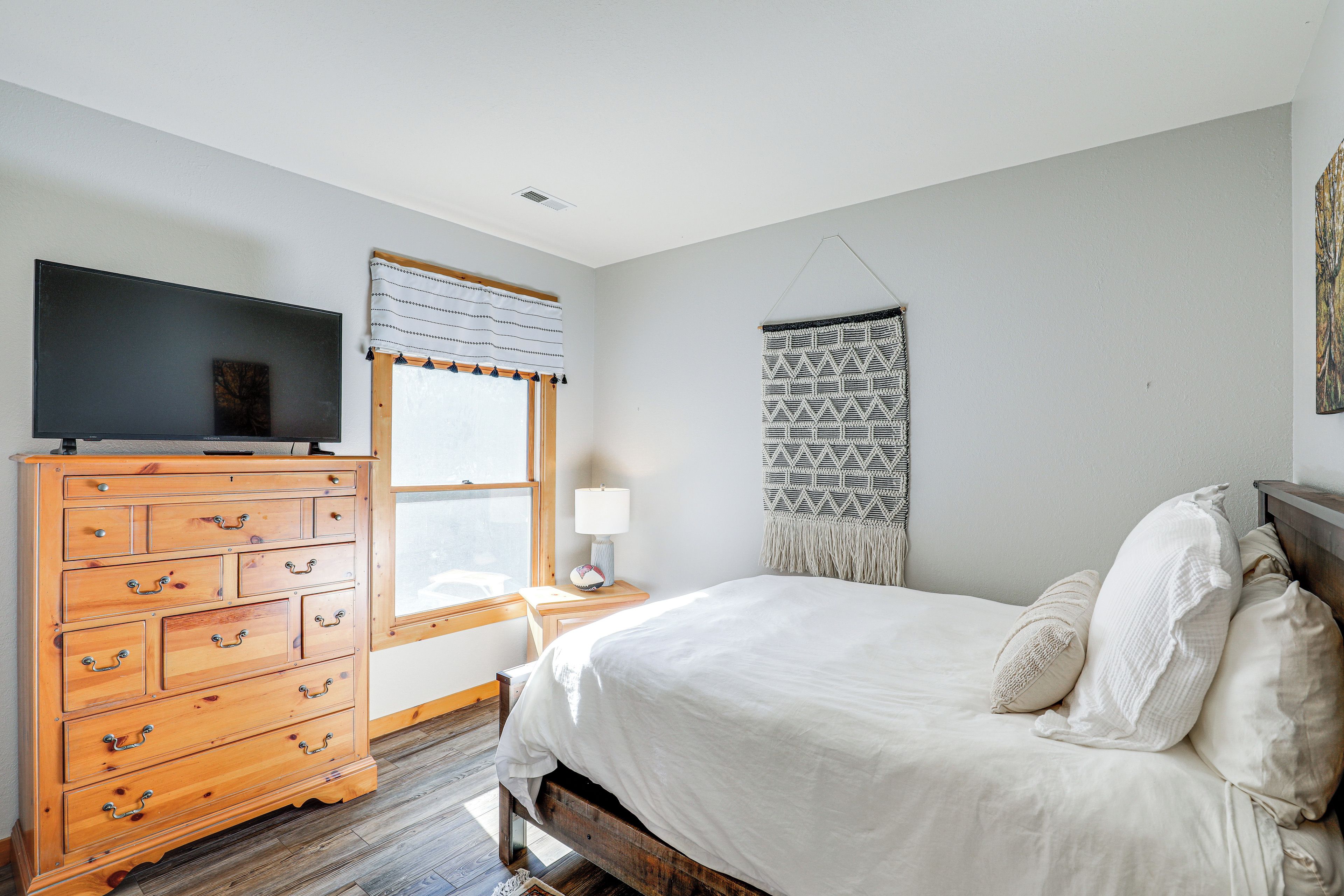 A bedroom with a wooden dresser, a bed with white linens, a wall-mounted TV, and a window with blinds.