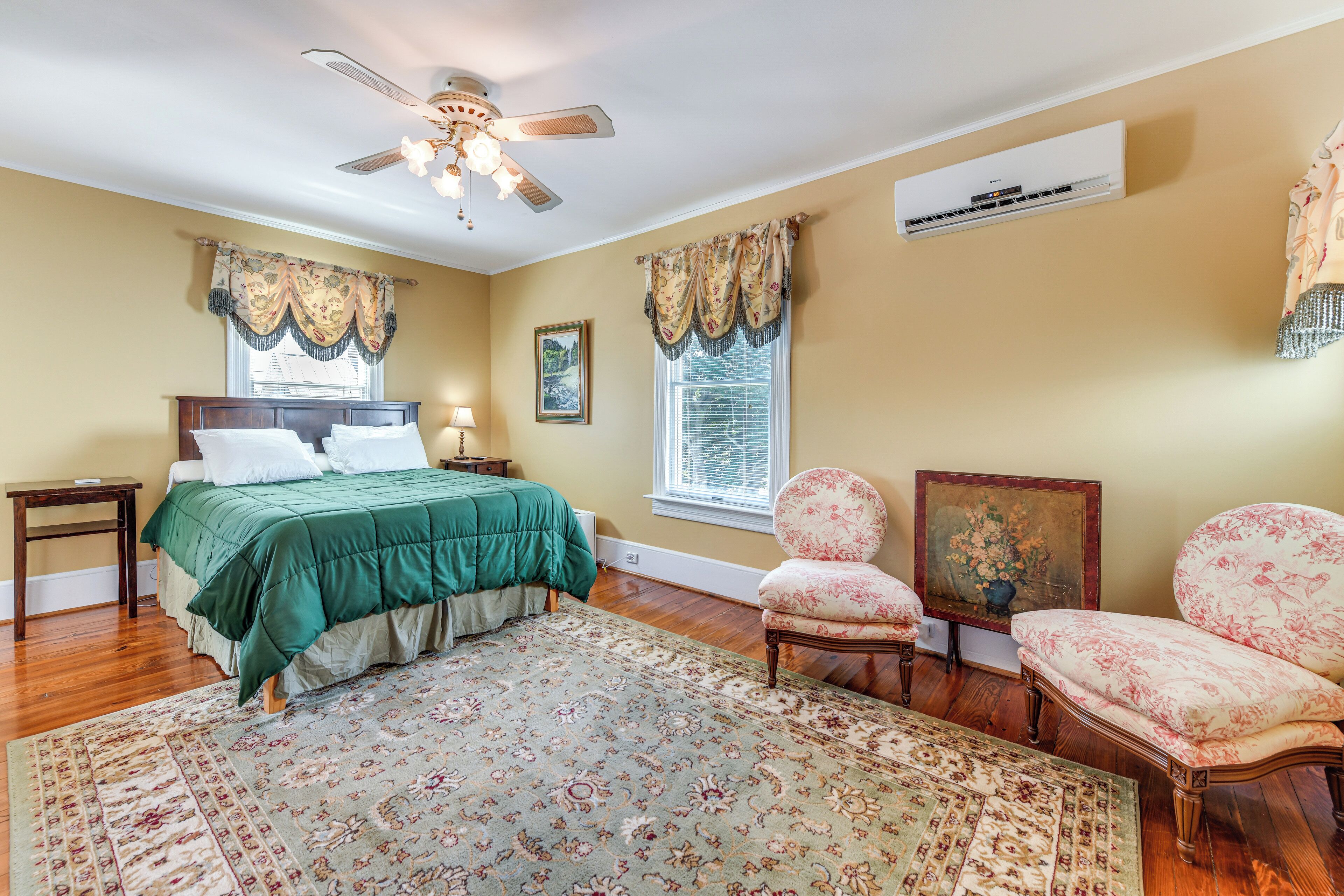 Un dormitorio con cama, dos mesitas de noche, una silla, alfombra, ventilador de techo y aire acondicionado.