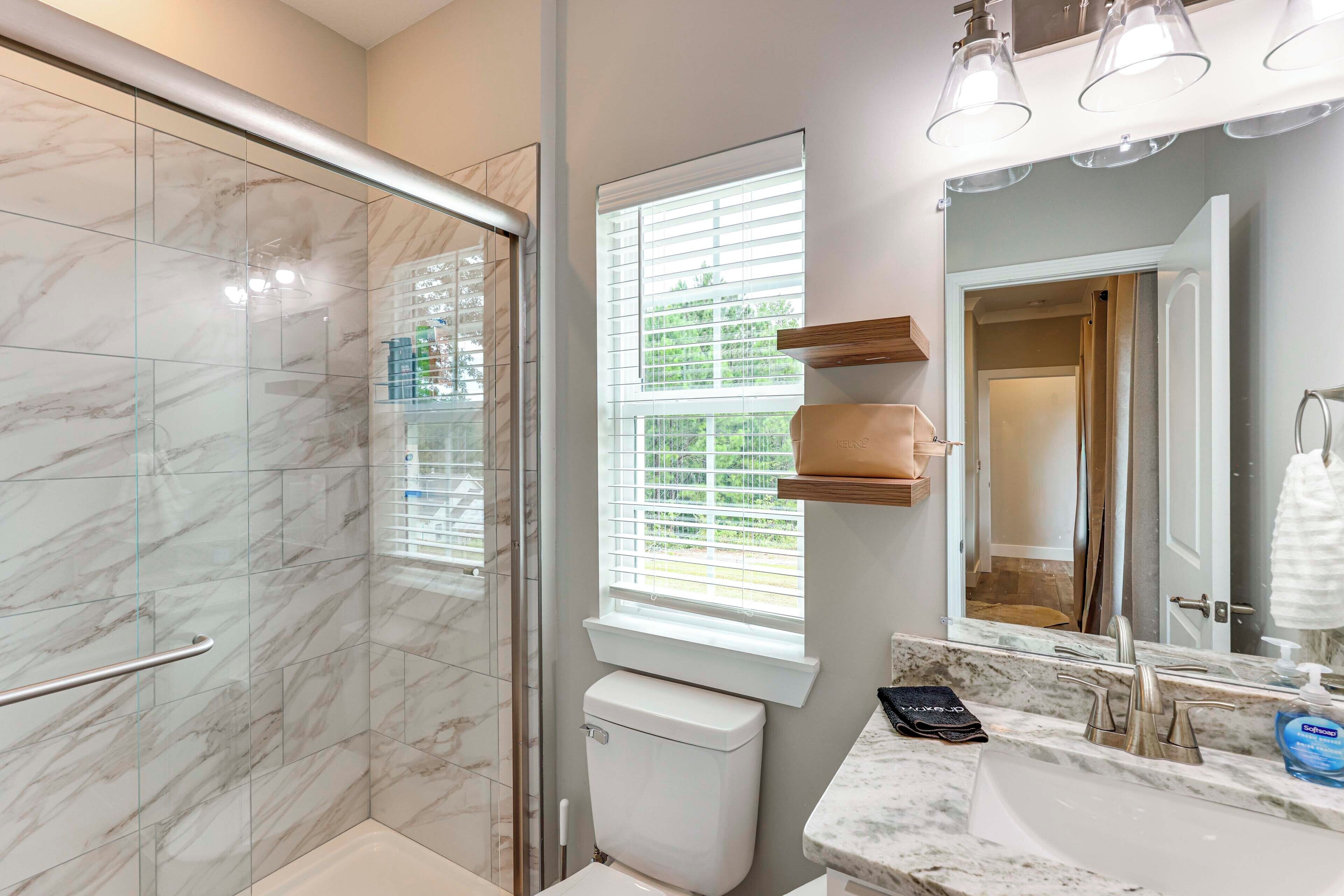 A modern bathroom with a glass shower enclosure, a toilet, a sink with a marble countertop, and a window with blinds.
