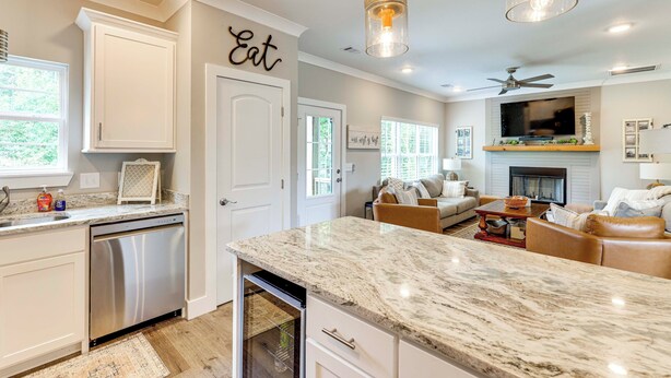 A modern kitchen with white cabinets, a dishwasher, and a marble countertop. There is a dining area with a sofa and a fireplace. - Walk to Lake Martin: Water-view Retreat w/ Deck! (Tallassee)