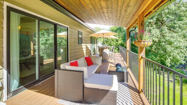 A covered outdoor seating area with wicker furniture, a glass door leading to a patio, and a wooden railing overlooking a garden. - Modern Creekside Retreat 1 Mi to Spirit Mtn Casino (Grand Ronde)