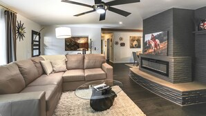 A modern living room with a sectional sofa, a glass coffee table, a fireplace, and a ceiling fan. - Modern Creekside Retreat 1 Mi to Spirit Mtn Casino (Grand Ronde)