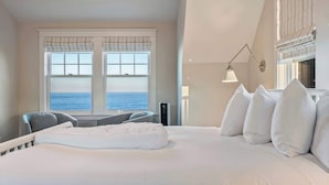 A bedroom with a large bed, a view of the sea through the windows, and a seating area with a chair and table. - Oceanfront Home w/ Semi-Private Beach • Sleeps 12 (York)