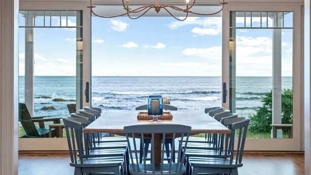 A dining area with a view of the ocean, featuring a chandelier, a long table, and multiple chairs. - Oceanfront Home w/ Semi-Private Beach • Sleeps 12 (York)