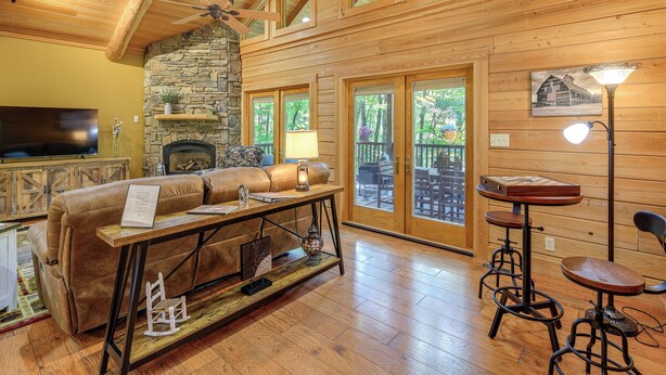 A cozy cabin interior with a stone fireplace, wooden ceiling, and a large window overlooking a deck. - Luxe Mountain Escape w/ Hot Tub & Gym Near Boone! (Zionville)