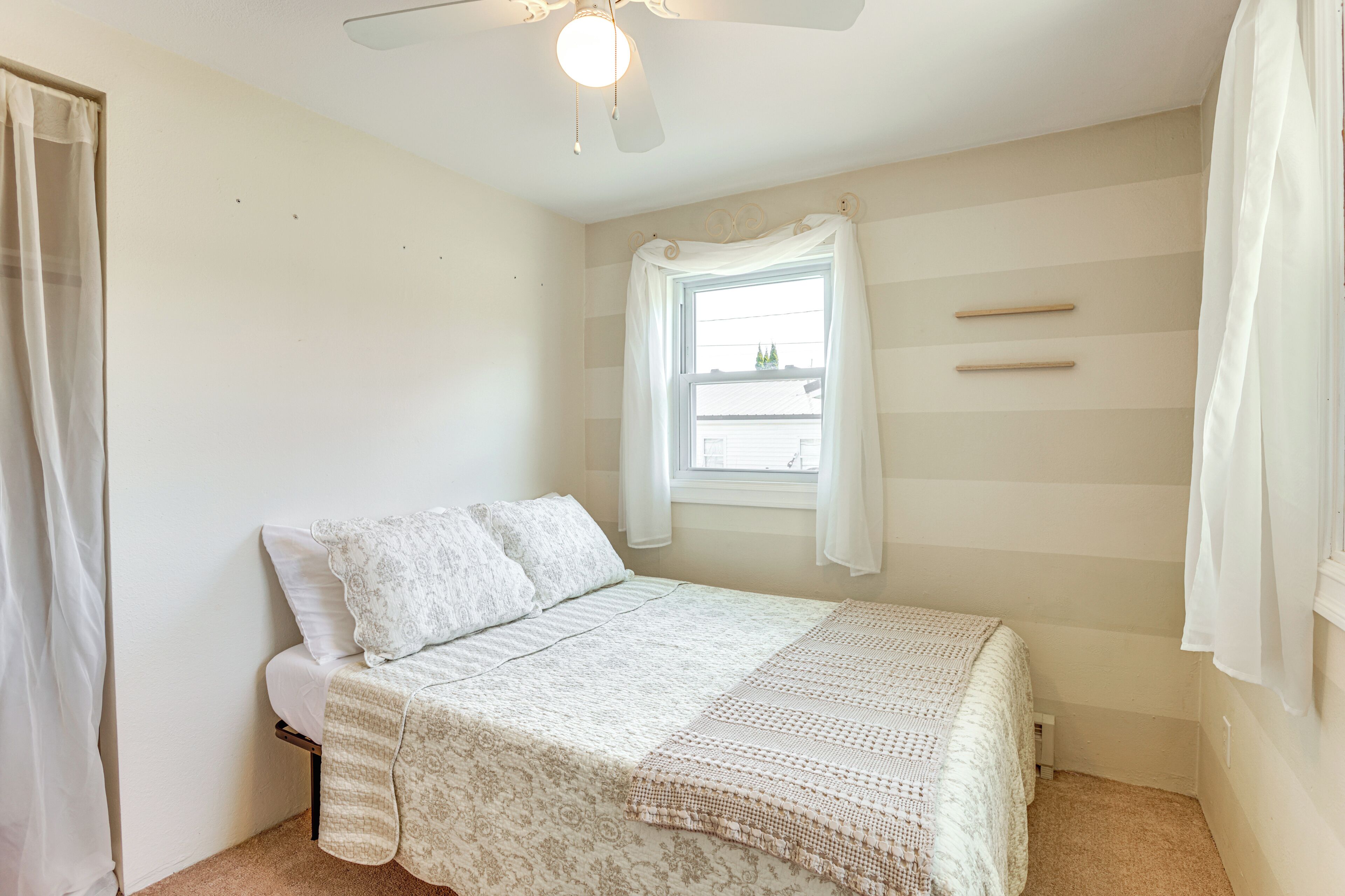 A bedroom with a bed, a window with curtains, a ceiling fan, and a door.