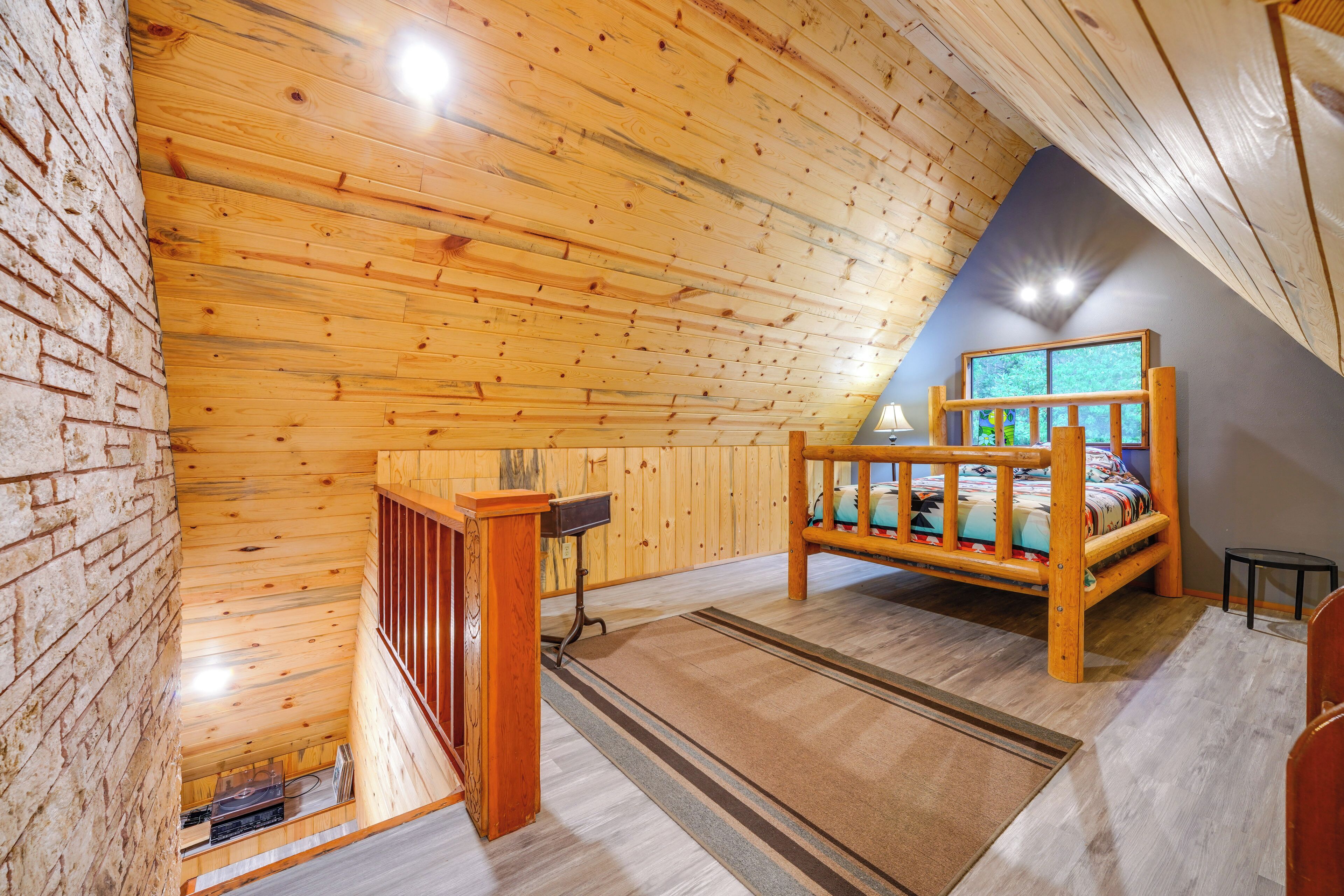 Quarto em estilo loft com teto de ripas de madeira, uma cama, uma mesinha e janela com vista para o verde.