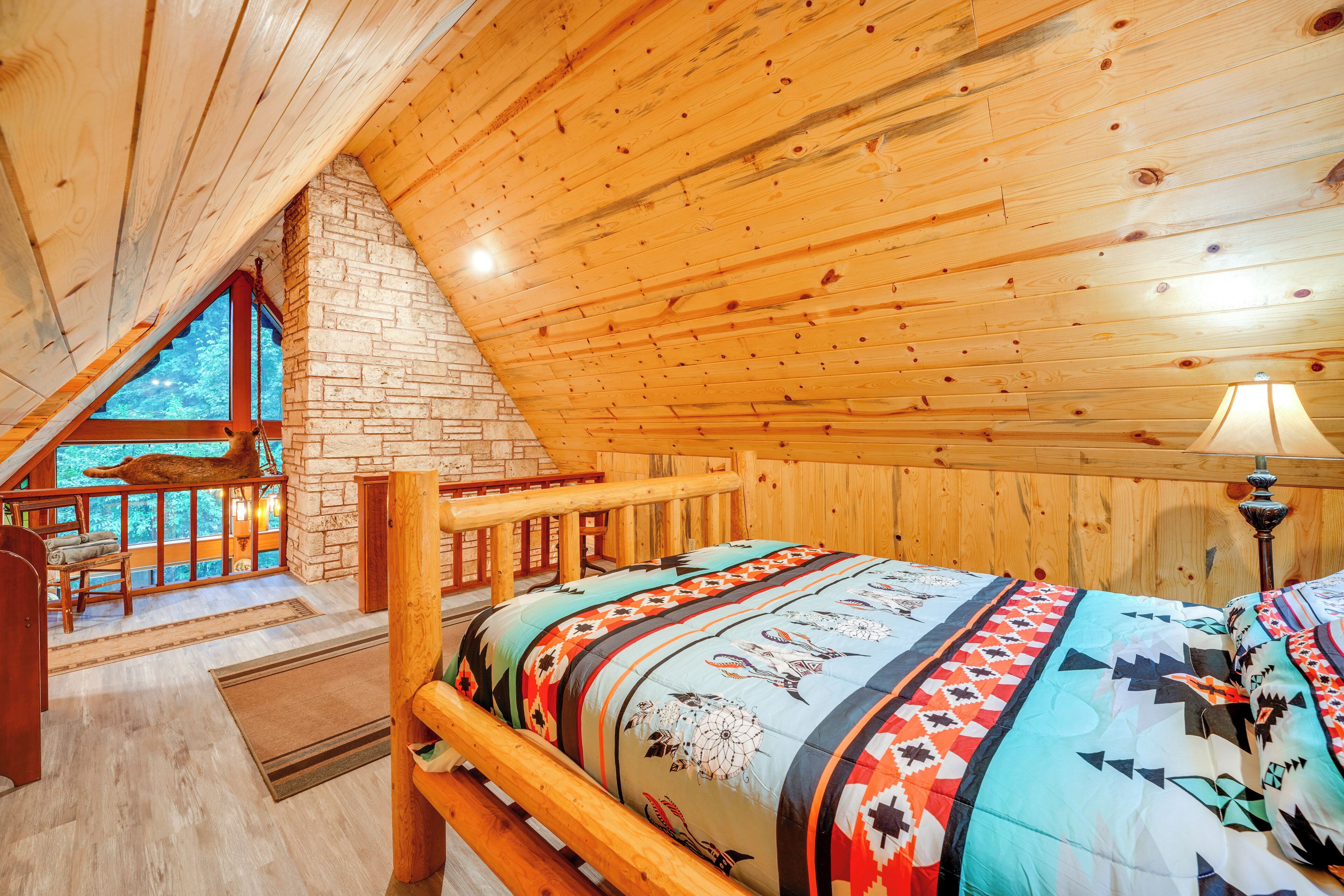 A cozy bedroom with a wooden ceiling, a stone fireplace, a large window, and a bed with a colorful quilt.