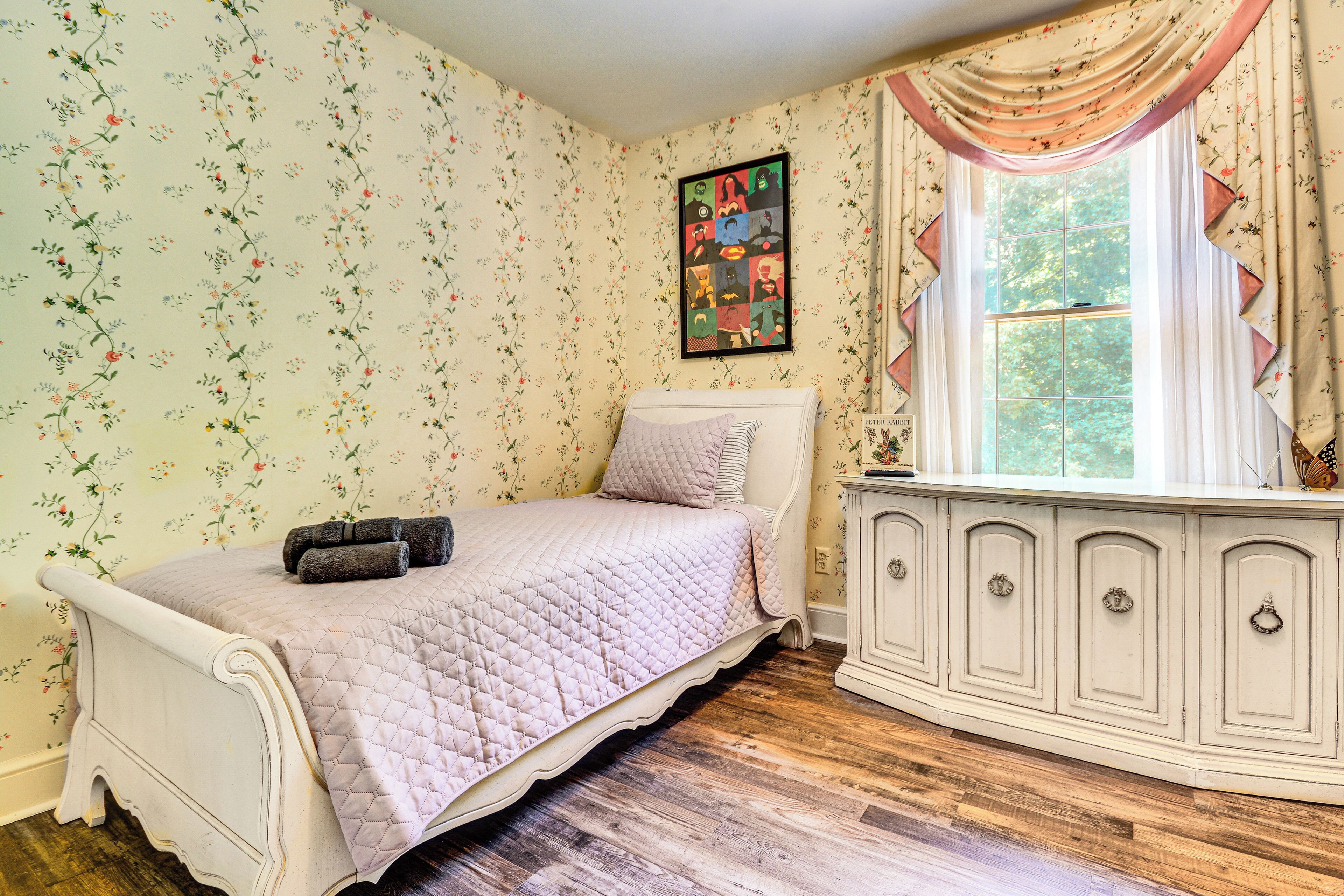 A bedroom with floral wallpaper, a bed with a quilted pink bedspread, a dresser with multiple drawers, and a window with curtains.