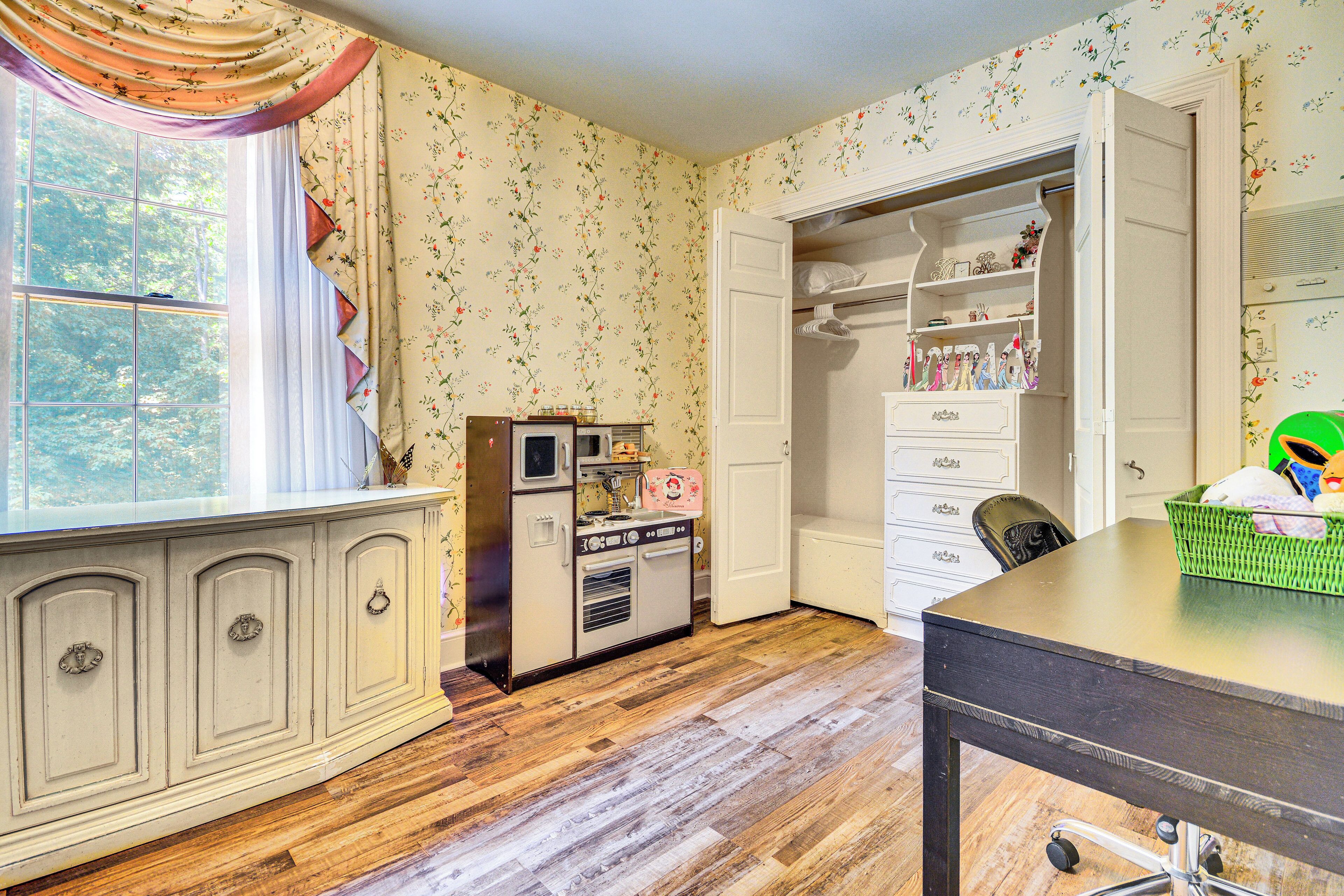 Une pièce dotée d’une kitchenette au style rétro, d’un plancher en bois, d’une fenêtre avec des rideaux et d’un placard où sont suspendus des vêtements.