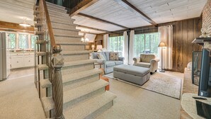 A spacious living area with a wooden staircase, a fireplace, and a large window offering a view of the outdoors. - Cabin Near Lake w/ Deck, Fire Pit & Grill! (Elmira)