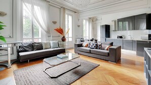 Een ruime woonkamer met een grijze bank, een glazen salontafel en een groot raam met dunne gordijnen.