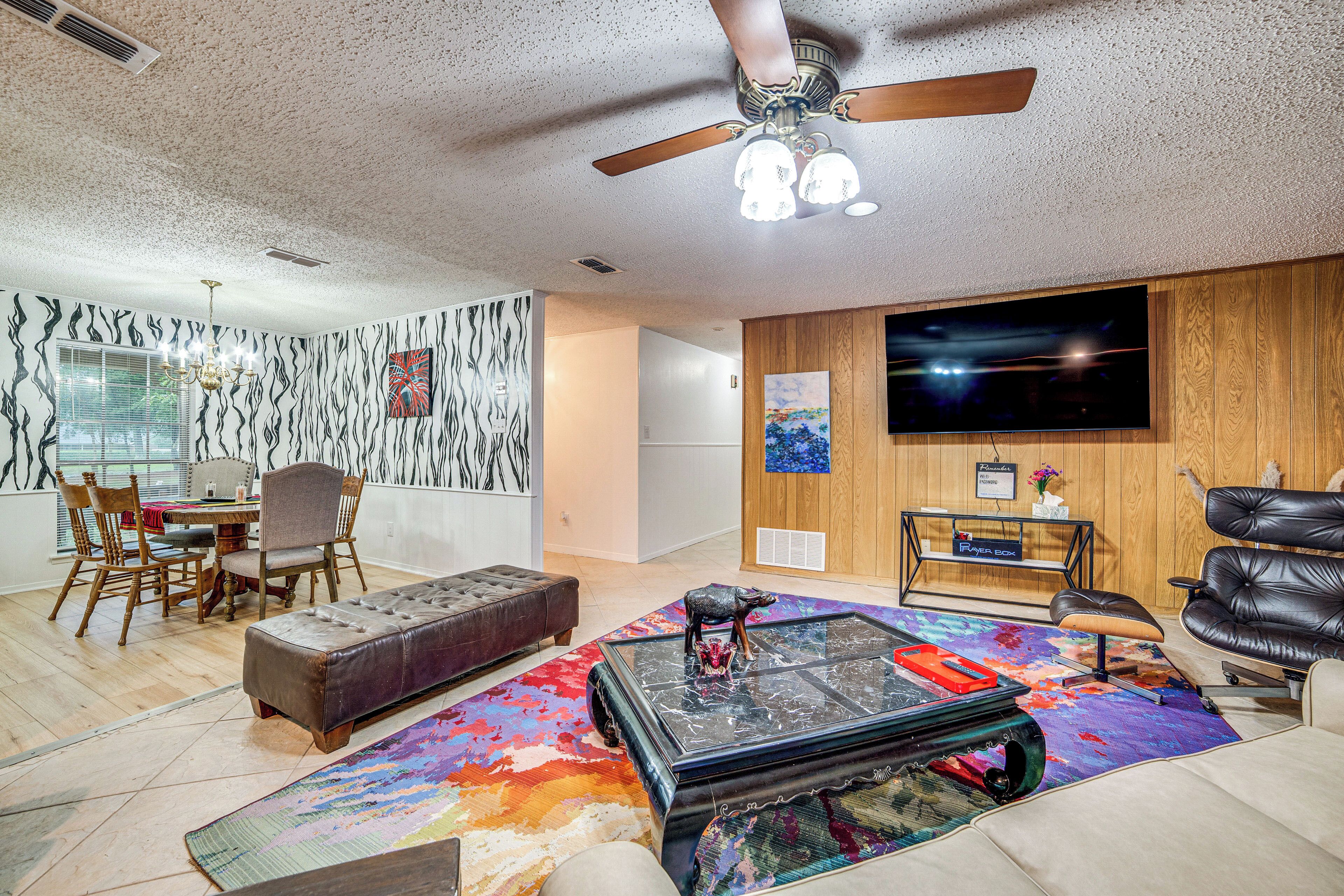 A modern living room with a ceiling fan, a television, a dining area with chairs, and a colorful rug.