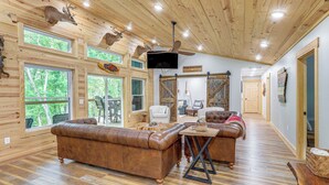 A spacious cabin interior with a wooden ceiling, large windows, a brown leather sofa, a television, and a sign reading "White River Cabin". - Relax on the River: Modern Mountain View Escape! (Mountain View)