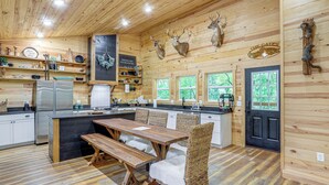 A rustic kitchen with wooden walls, a central island, and a dining area with wicker chairs. - Relax on the River: Modern Mountain View Escape! (Mountain View)