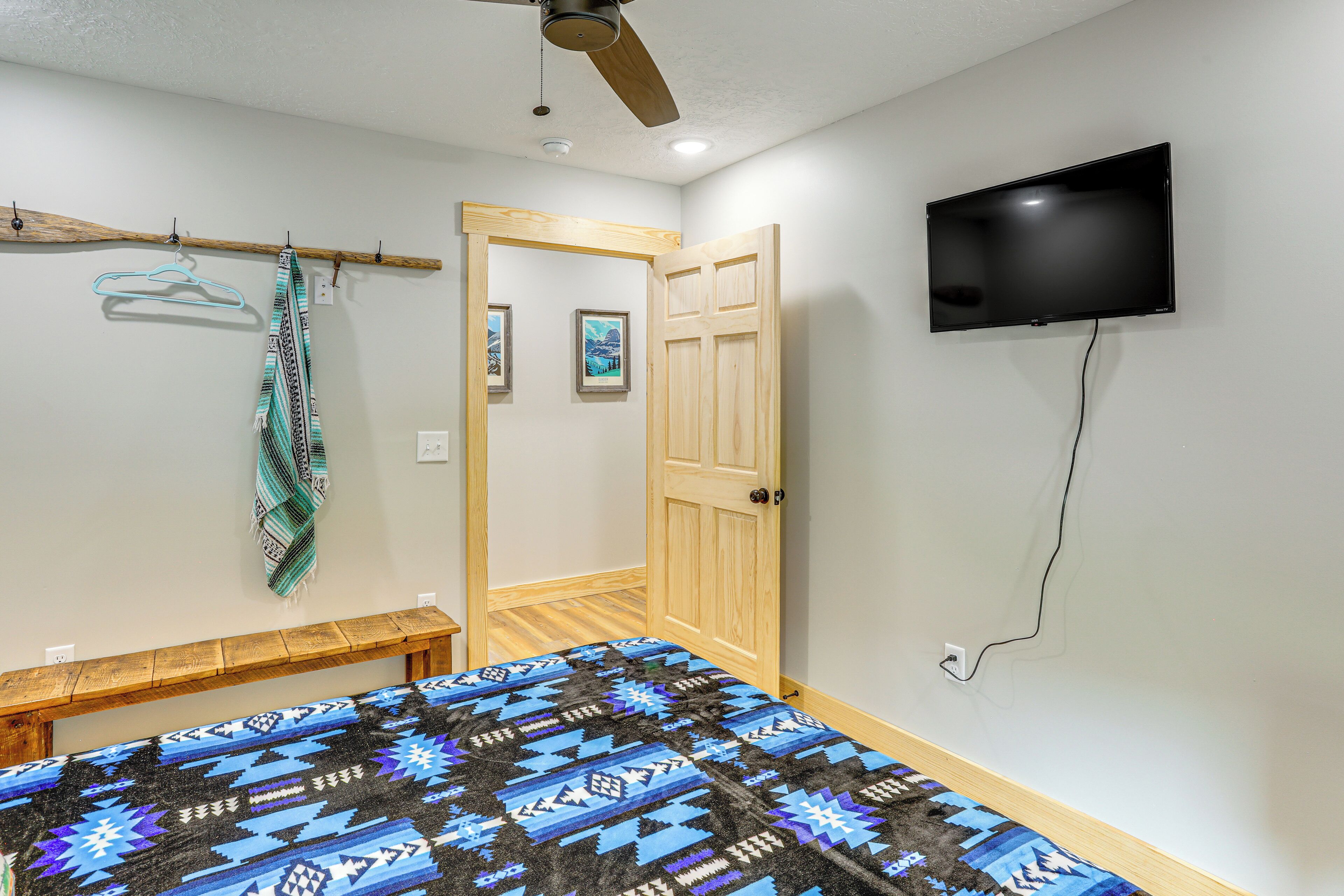 A bedroom with a bed, a wooden bench, a ceiling fan, a TV mounted on the wall, and a door leading to another room.