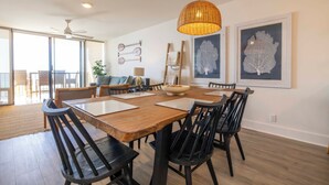 Een eethoek met een houten tafel, zwarte stoelen en een geweven hanglamp. Er hangen kunstwerken in lijsten aan de muur en er is een plafondventilator.
