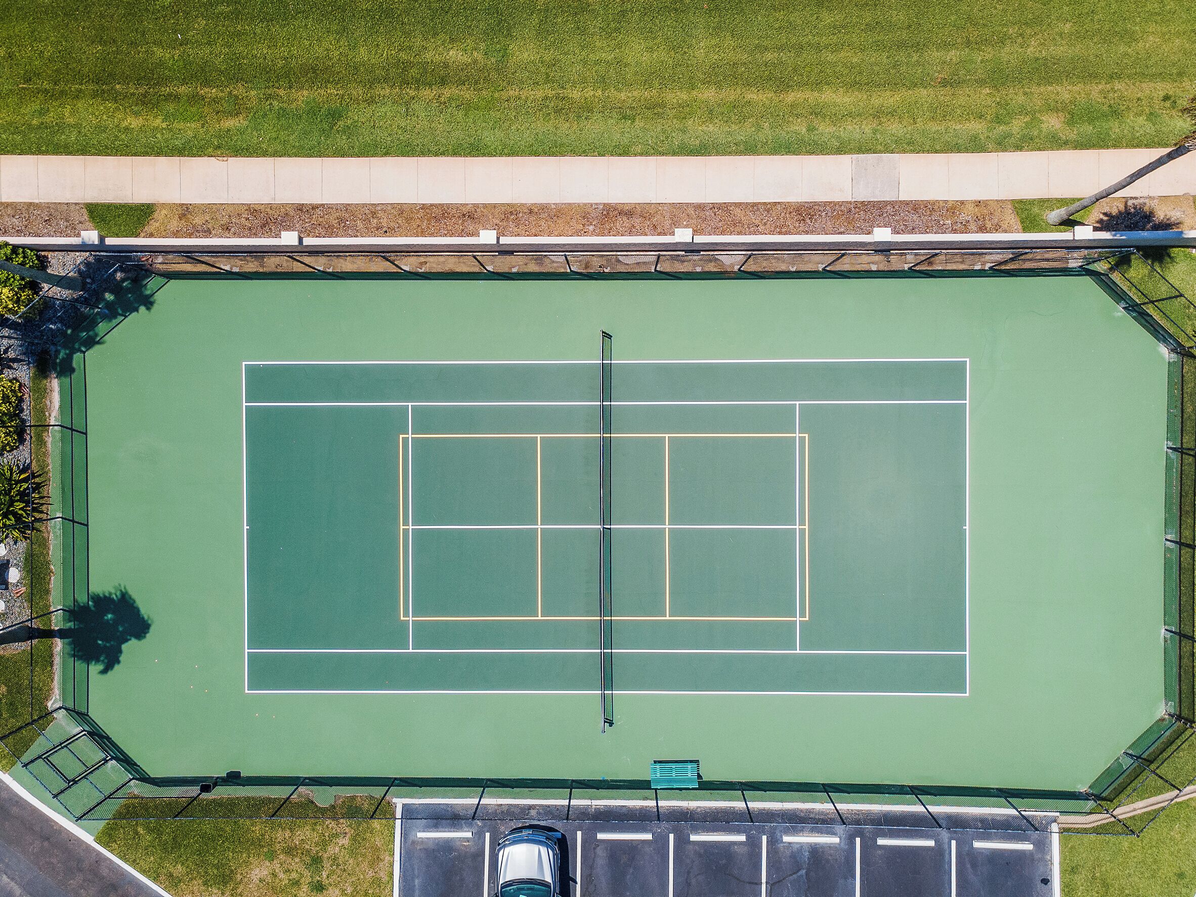 Vue aérienne d’un court de tennis entouré d’une clôture, avec un espace de stationnement et un petit banc vert.