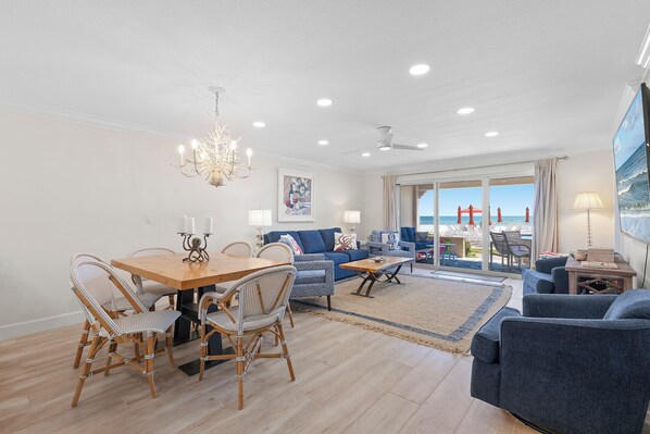 A spacious living area with a dining table, chairs, sofas, and a view of the beach through large windows.