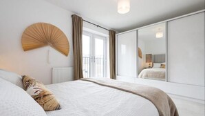 A modern bedroom with a large bed, a wall fan, a window with curtains, and a mirror reflecting the room.