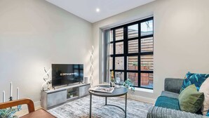 Ein modernes Wohnzimmer mit Flachbildfernseher, rundem Couchtisch, einer Couch und einem großen Fenster mit Blick ins Freie.