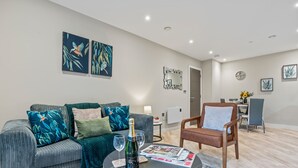 Ein modernes Wohnzimmer mit einem grauen Sofa, einem Holzstuhl, einem Couchtisch mit Flasche und Gläsern sowie Kunstwerken an den Wänden.