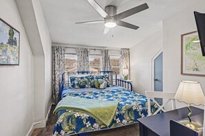 Ein Schlafzimmer mit einem Bett, einem Deckenventilator, einem Schreibtisch und einem Fenster mit Vorhängen.