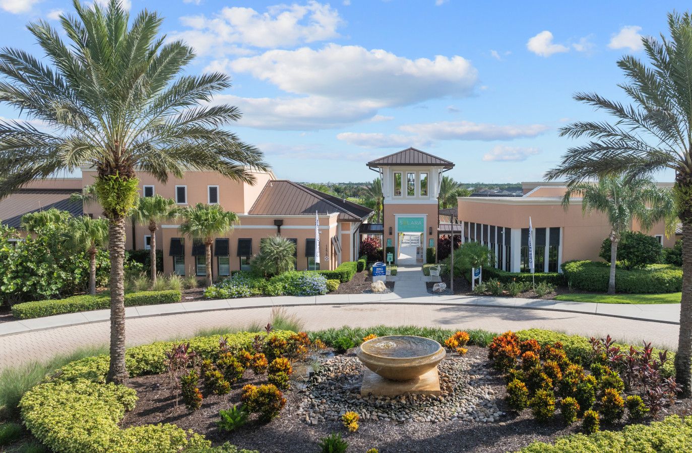A hotel entrance with a central tower, palm trees, and a landscaped garden with a water feature.