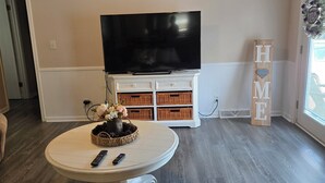 Sala de estar con televisor de pantalla plana sobre un mueble blanco, una mesa redonda de centro con una cesta y controles remotos, y un letrero que dice 'HOME'.